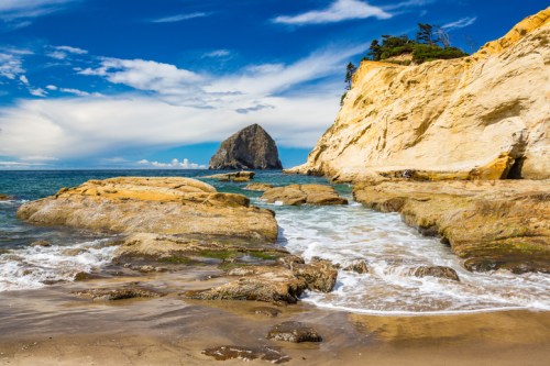 Scenic beach view with rocky shore, gentle waves, and a prominent sea stack under a blue sky with clouds.