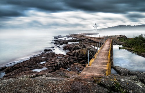 A foggy scene of a rocky pier leading to a lighthouse, surrounded by calm waters and overcast skies.