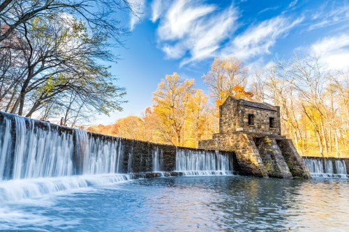A scenic view of a stone structure beside a waterfall, surrounded by autumn trees and a clear blue sky.