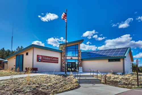 A modern visitor center with solar panels, surrounded by grass and under a blue sky with clouds. An American flag is flying.
