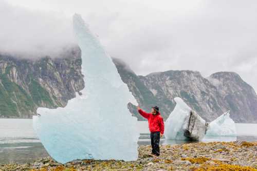 A person in a red jacket touches a large iceberg near a rocky shore, with mountains and cloudy skies in the background.