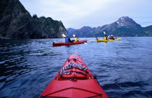 A red kayak in the foreground with two kayakers in yellow and blue paddling in a scenic mountain landscape.