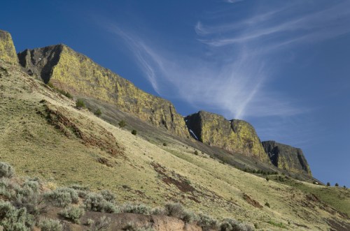 Lush green hills lead up to rugged, moss-covered cliffs under a blue sky with wispy clouds.