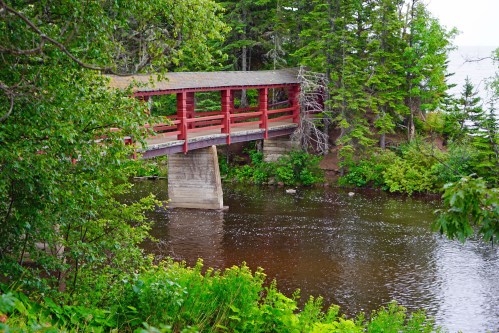 A red wooden bridge spans a calm river, surrounded by lush green trees and foliage.