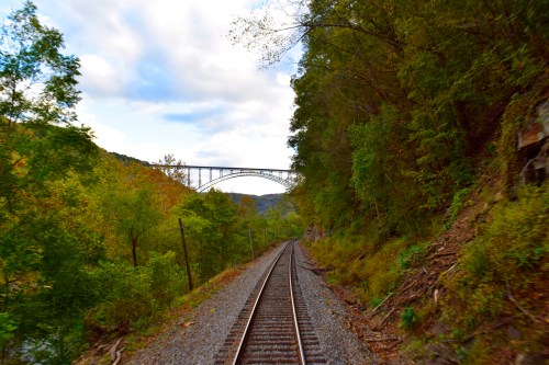 A view of train tracks leading towards a bridge, surrounded by lush green trees and a cloudy sky.