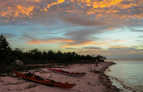 Two kayaks on a sandy beach at sunset, with colorful clouds and a calm sea in the background.