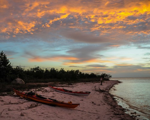 Two kayaks on a sandy beach at sunset, with vibrant clouds and calm water in the background.