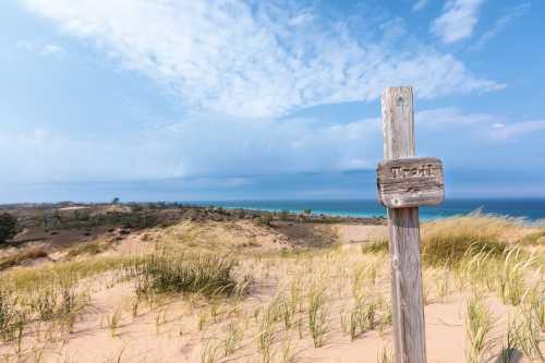 A wooden trail sign stands in a sandy landscape with grass, overlooking a blue lake and cloudy sky.