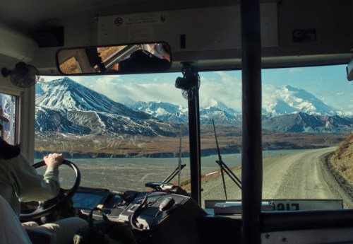 View from a bus driver's seat, showcasing mountains and a gravel road against a clear blue sky.