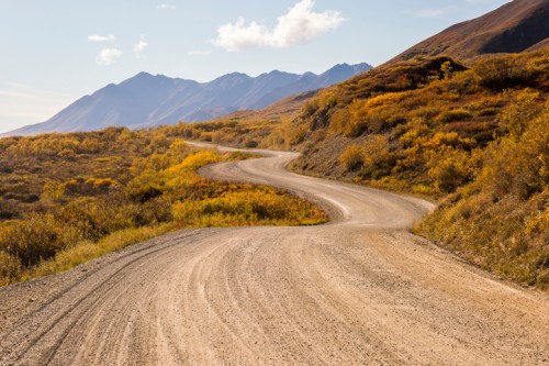 A winding dirt road through colorful autumn foliage with mountains in the background under a clear blue sky.