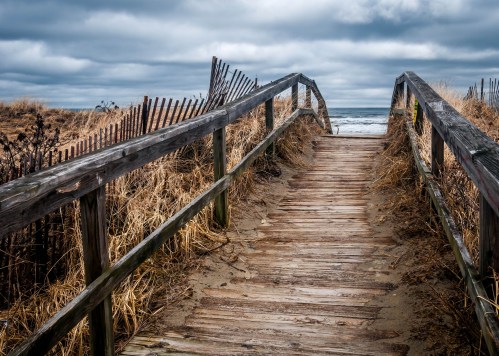 A wooden walkway leads to a beach, surrounded by tall grass and a cloudy sky.