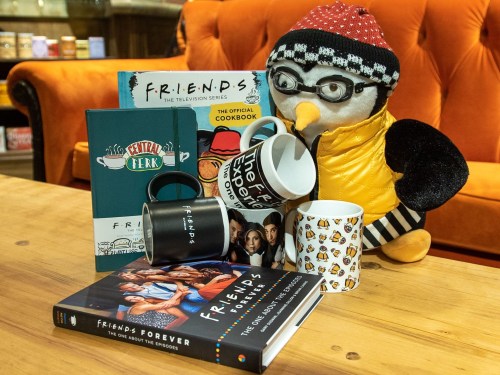 A penguin plush toy surrounded by "Friends" merchandise, including mugs and cookbooks, on a wooden table.