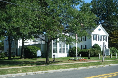 A white house with large windows surrounded by trees and a well-maintained lawn, located along a road.