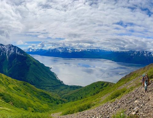 A panoramic view of a lush green mountain landscape with a lake and snow-capped mountains under a cloudy sky.