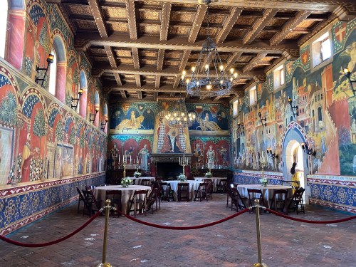 A vibrant, historic room with colorful murals, wooden beams, and tables set for an event, surrounded by elegant decor.