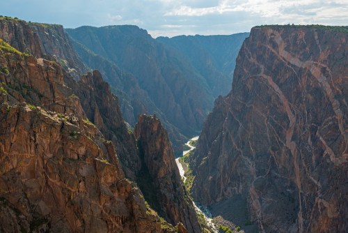 A breathtaking view of a deep canyon with steep rock walls and a winding river at the bottom, under a cloudy sky.