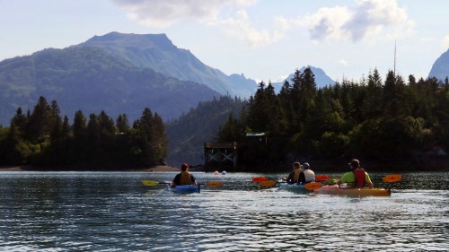 Four kayakers paddle on a calm lake surrounded by mountains and lush trees under a partly cloudy sky.