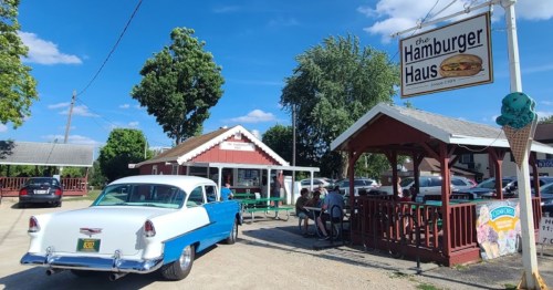A vintage blue car parked outside the Hamburger Haus, with people sitting at picnic tables under a clear blue sky.