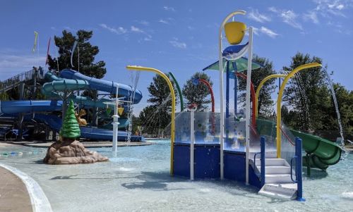 A water park scene featuring colorful slides, a splash pad, and trees under a clear blue sky.