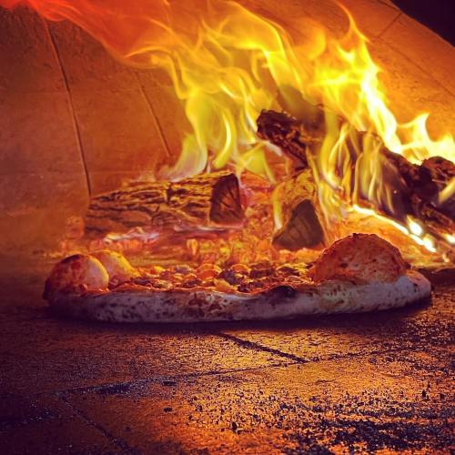 A pizza baking in a wood-fired oven, with flames and glowing embers in the background.