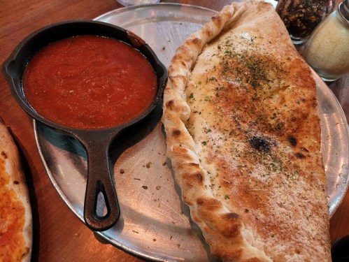 A golden-brown calzone on a metal tray, accompanied by a small cast-iron bowl of marinara sauce.