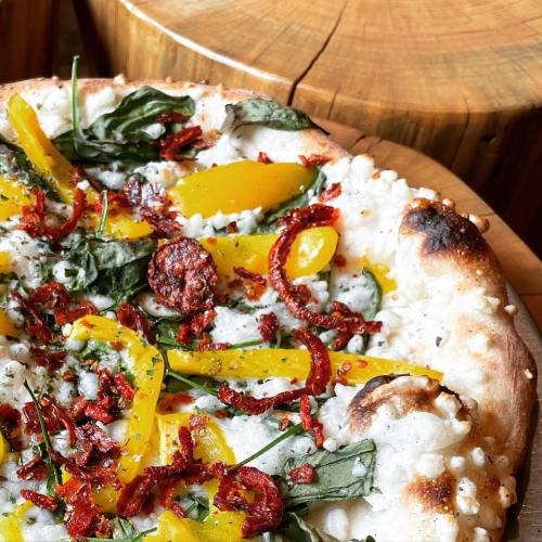A close-up of a pizza topped with yellow peppers, spinach, sun-dried tomatoes, and a creamy white sauce on a crust.