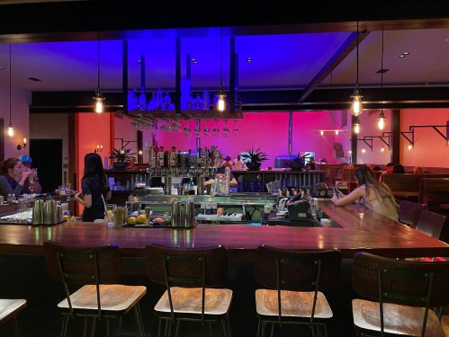 A dimly lit bar with colorful lighting, featuring a wooden counter and patrons seated at the bar.