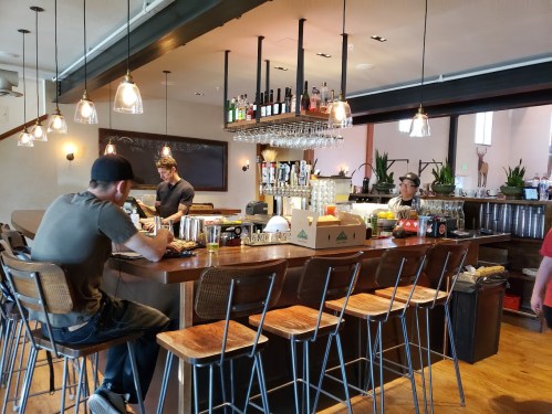 A modern bar with patrons seated at the counter, bottles on display, and a bartender preparing drinks.