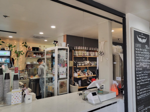 A cozy café interior with a counter, shelves of products, and a barista preparing drinks in the background.