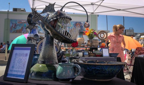 A unique ceramic fish sculpture with a wide mouth, displayed at an outdoor market with colorful pottery and a vendor in the background.