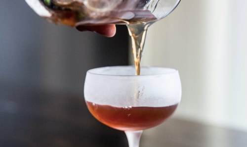 A cocktail being poured from a glass pitcher into a chilled coupe glass.