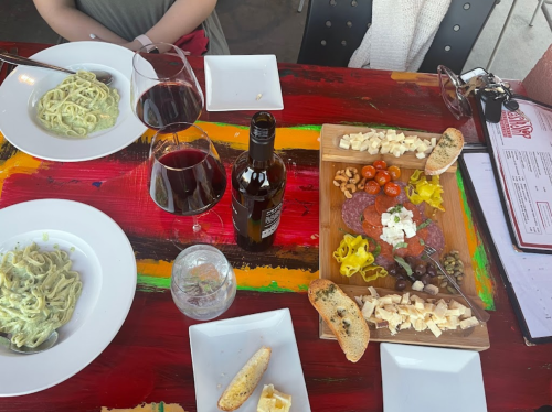 A colorful table with two plates of pasta, a charcuterie board, wine glasses, and a bottle of wine.