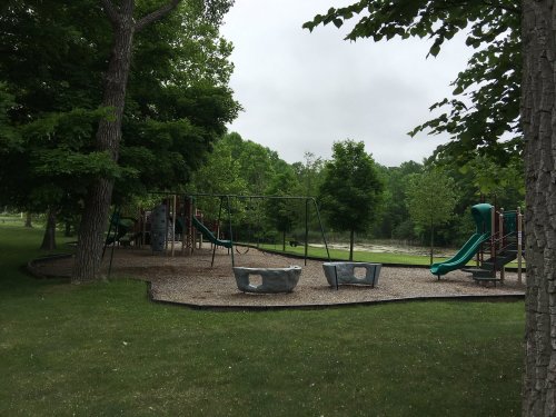 A playground with swings, a slide, and climbing structures, surrounded by trees and grassy areas on a cloudy day.