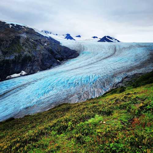 A vast glacier flows down a mountain, surrounded by green hills and rocky peaks under a cloudy sky.