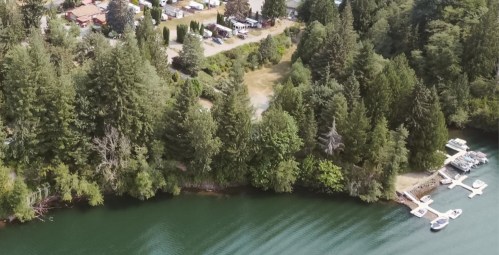 Aerial view of a serene riverbank with boats docked, surrounded by lush green trees and a campsite in the background.