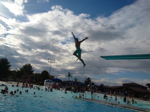 A person jumps off a diving board into a crowded pool on a sunny day, with clouds in the background.