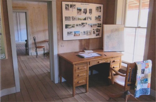 A wooden desk with a chair, surrounded by light-colored walls and a poster of images, in a cozy, rustic room.