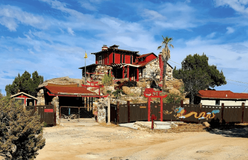 A unique stone and wood house with red accents, surrounded by desert landscape and decorative elements, including a dragon mural.