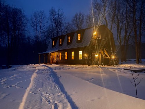 A cozy cabin in the snow at night, with warm lights glowing from the windows and trees in the background.