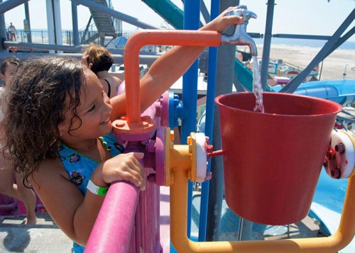 A child joyfully pours water from a colorful faucet into a red bucket at a water park.