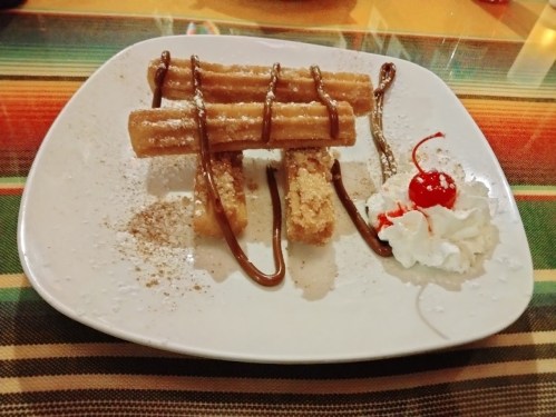 A plate of churros drizzled with chocolate, topped with whipped cream and a cherry, served on a colorful tablecloth.