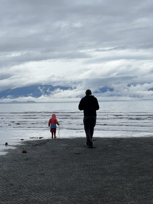 A person and a child in raincoats walk along a beach with cloudy skies and distant mountains in the background.