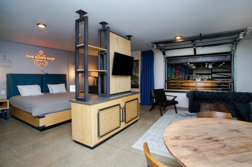 A modern hotel room featuring a bed, TV, seating area, and a garage door opening to a colorful space.