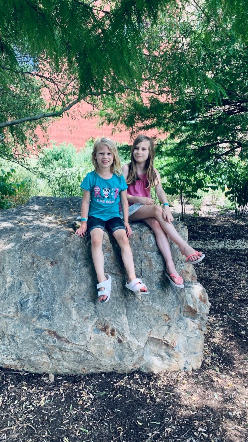 Two young girls sit on a large rock in a shaded area, smiling and enjoying a sunny day outdoors.