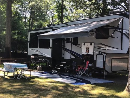 A parked RV with an awning, surrounded by trees, featuring a picnic table and chairs on a grassy area.