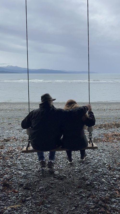 A couple sits on a swing by the beach, overlooking the water and distant mountains on a cloudy day.