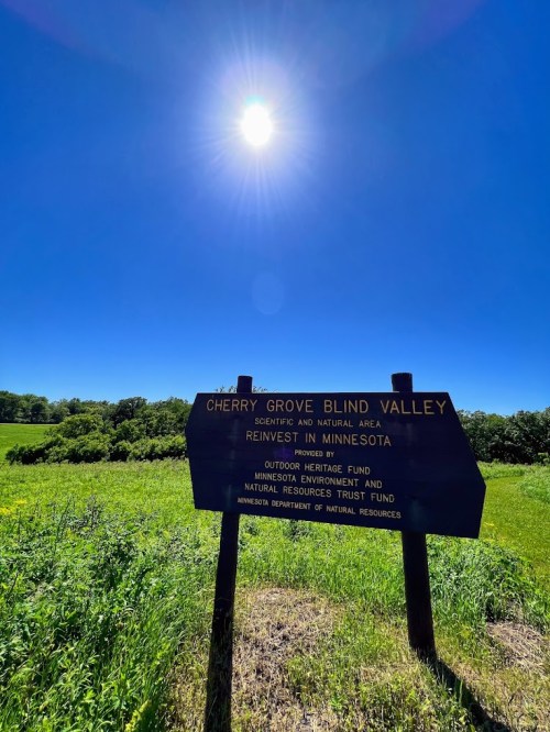Sign for Cherry Grove Blind Valley, a scenic area in Minnesota, with a bright sun overhead and green fields in the background.