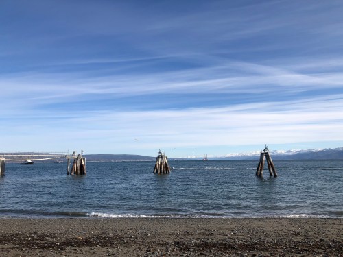 A serene coastal scene with wooden pilings in the water, mountains in the distance, and a clear blue sky.