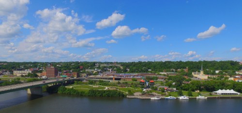 A scenic view of a riverfront city with green parks, buildings, and a blue sky filled with fluffy clouds.