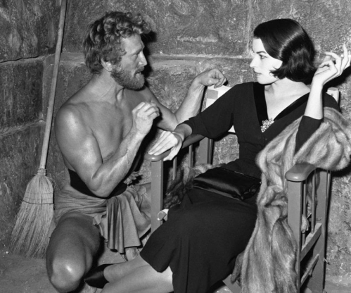 A shirtless man and a woman in a black dress sit in a dimly lit room, engaged in conversation.